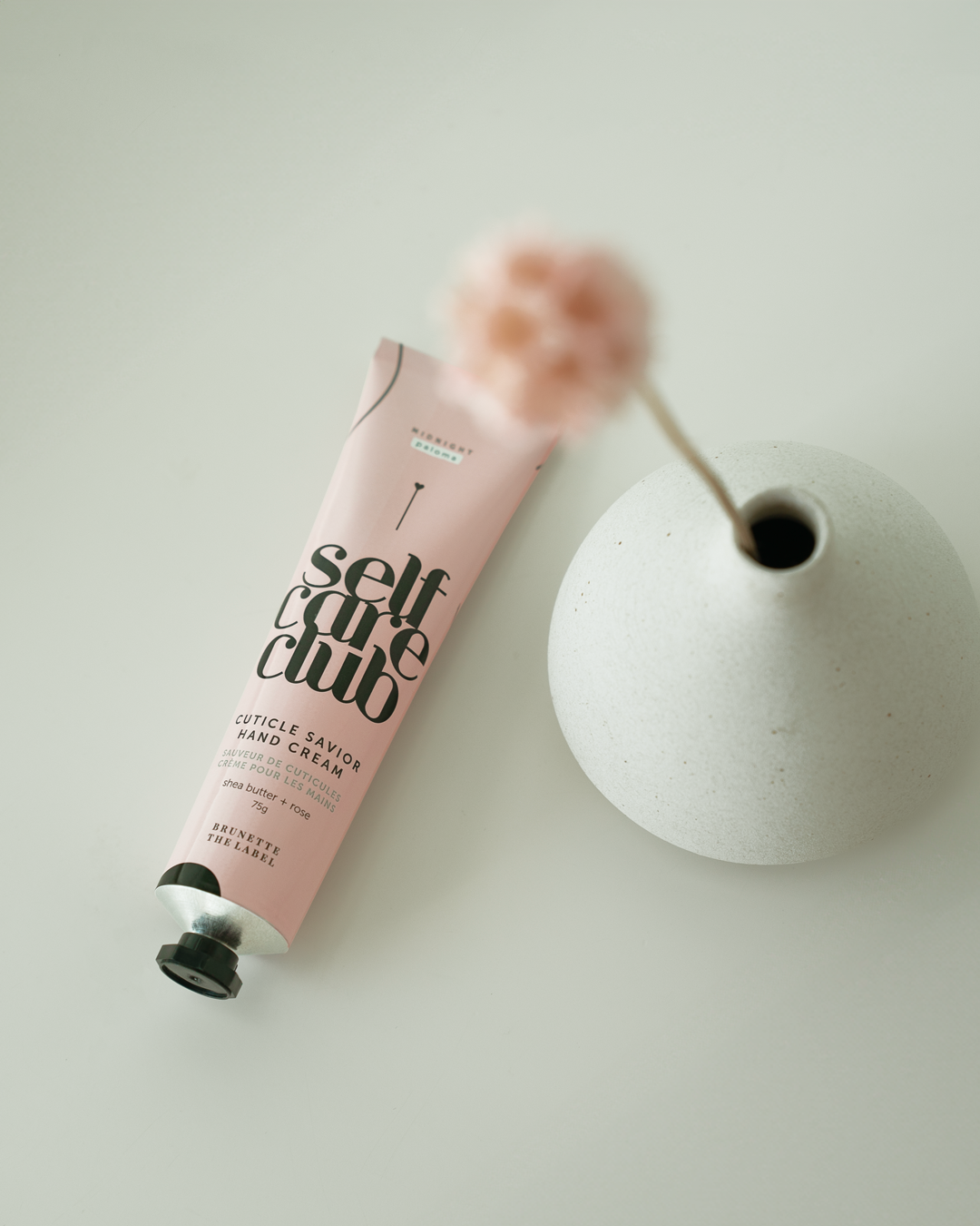 Hand cream tube labeled 'self care club' next to a white vase on a light surface
